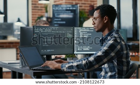 Portrait of african american developer using laptop to write code sitting at desk with multiple screens parsing algorithm in software agency. Coder working on user interface using portable computer. Royalty-Free Stock Photo #2108122673