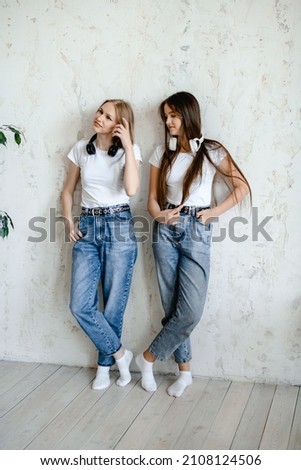 wo teenage girls stand near a white wall in funny sunglasses. they are wearing white t-shirts and blue jeans. headphones hang on their necks, they have fun, they indulge and laugh