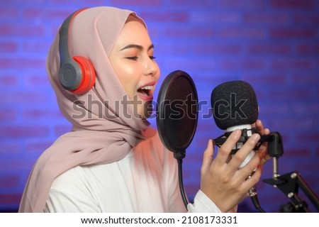 A young smiling muslim female singer wearing headphones with a microphone while recording song in a music studio with colorful lights.