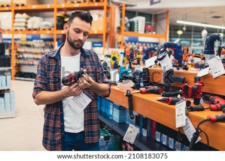 a man in a hardware store chooses a new screwdriver next to a showcase of power tools for repairs in the house Royalty-Free Stock Photo #2108183075