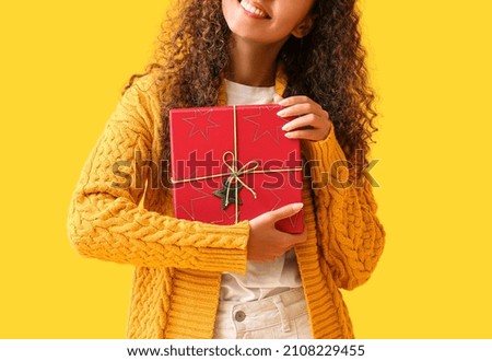 Young African-American woman in warm sweater with Christmas present on yellow background
