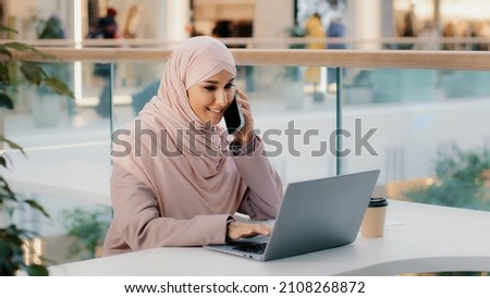 Arab multitasking woman sitting working on laptop talking on mobile telephone remotely consulting client using smartphone checks social network answering phone call writes notes makes online purchase