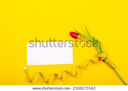 Tulip, white business card blank and centimeter tape on yellow background with copy space, text place. Fitness greeting card. International holiday. Sewing handicraft hobby store. Slimming certificate