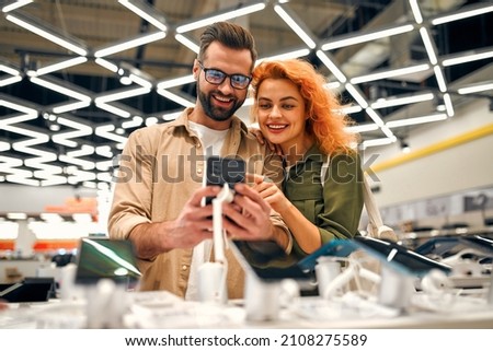 Red-haired sweet woman with her boyfriend chooses new smartphone in store of household appliances, electronics and gadgets. Royalty-Free Stock Photo #2108275589