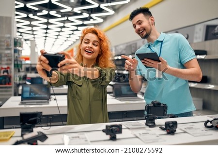 Young woman receives advice from seller in store of household appliances and gadgets, buying camera. Male consultant helps in choosing new gadget.