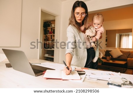 Female interior designer making notes while holding her baby. Multitasking mom planning a new project in her home office. Creative businesswoman balancing work and motherhood.