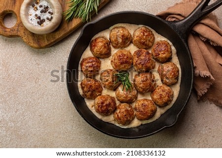 Homemade Swedish Meatballs made with ground meat, onion, egg, bread crumbs and nutmeg. With creamy gravy in black pan skillet.  On beige concrete table. Royalty-Free Stock Photo #2108336132