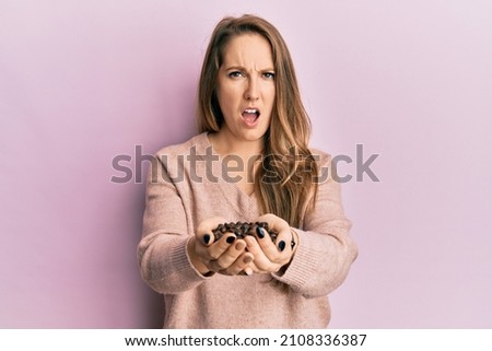 Young blonde woman holding coffee beans with hands in shock face, looking skeptical and sarcastic, surprised with open mouth 