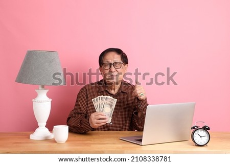 The senior Asian man sitting at working desk with laptop on the pink background.
