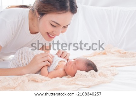 Selective focus of Asian young mother swaddling baby on bed, Beautiful woman wrapping adorable infant in thin cloth. Cute newborn child taking nap after bath. Concept of baby care and family. Royalty-Free Stock Photo #2108340257