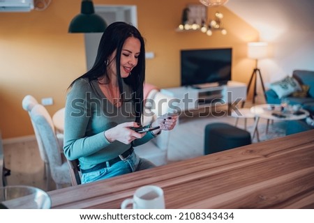 Middle aged woman holding bank credit card in hands and enjoying shopping online in a mobile app while sitting at a kitchen counter at home. Online purchase concept.