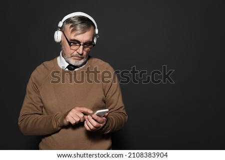 senior man in beige jumper and headphones pointing at mobile phone on dark grey