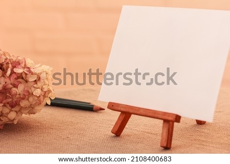 Wooden mini Easel with Blank White Canvas, paper, pencils, hydrangea, Hortensia on the table. Mock up for Artists and Painters. Mini Wooden Stand with Clean Artboard. Copy Space