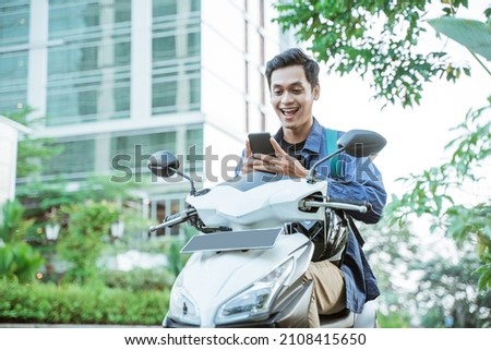 Smiling asian man using a cellphone while riding a motorcycle Royalty-Free Stock Photo #2108415650