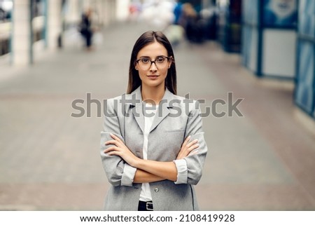 A young serious, elegant businesswoman standing on the street with arms crossed and looking at the camera. Success coming with hard work. A young proud woman on a street Royalty-Free Stock Photo #2108419928
