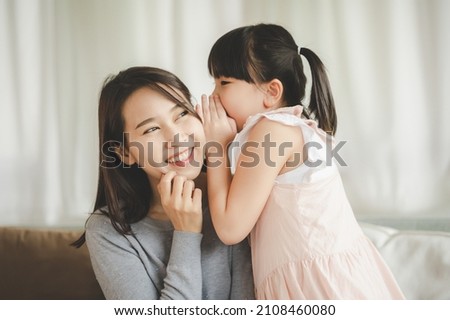 Little Asian daughter girl whispering I love you mommy to happy smiling mother in living room