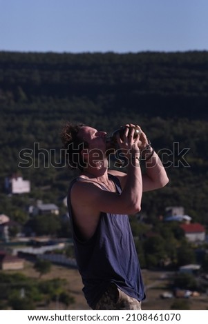 A curly guy with black hair and a red beard, dressed in a blue T-shirt and khaki pants, stands on a height against the backdrop of the hills and the city below them, closes a green army flask