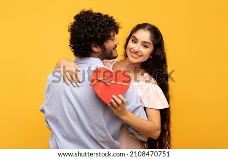 Young indian lovers hugging on yellow background, lady holding heart shaped gift box and smiling at camera. Loving couple celebrating Valentine's Day or anniversary together Royalty-Free Stock Photo #2108470751