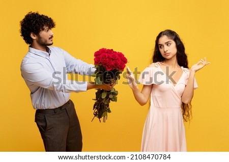 Unwanted confession. Displeased indian lady rejecting her admirer with roses on Valentine's Day, man giving flowers to his girlfriend, yellow studio background Royalty-Free Stock Photo #2108470784