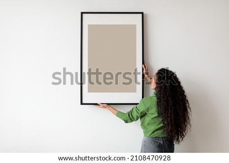 Happy Woman Hanging Empty Poster In Frame On White Wall Standing Indoors. Modern Art, Domestic Interior And Home Decor Pictures Concept. Mockup For Advertisement Royalty-Free Stock Photo #2108470928