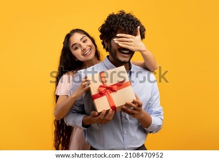 Pretty indian woman covering her boyfriend's eyes, holding wrapped gift box and greeting him with birthday or anniversary, standing over yellow studio background Royalty-Free Stock Photo #2108470952