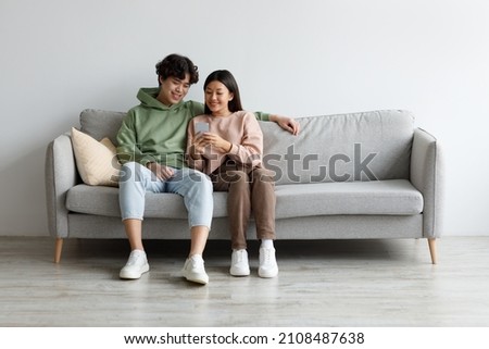 Happy young Asian couple using cellphone, browsing internet, chatting online, relaxing on sofa at home, free space. Cheery millennial family checking new mobile application, resting on couch together Royalty-Free Stock Photo #2108487638
