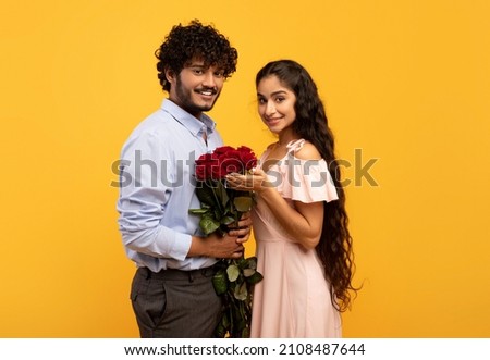 Lovers holiday. Romantic indian couple holding bouquet of flowers and looking at camera on yellow studio background. Loving guy pampering his girlfriend with roses on Valentine's Day