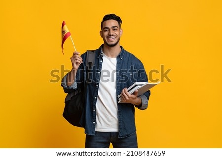 Positive young middle eastern man in casual with backpack and bunch of books student showing flag of Spain and smiling at camera over yellow studio background. Education abroad concept Royalty-Free Stock Photo #2108487659