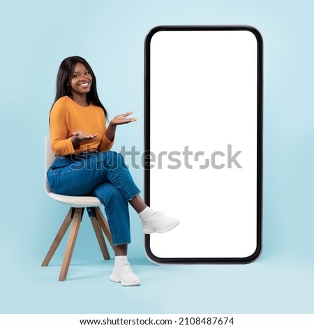 Full length of pretty black lady sitting on chair next to big blank smartphone with mockup for mobile app or website design, blue studio background, showing copy space for your online advertisement Royalty-Free Stock Photo #2108487674