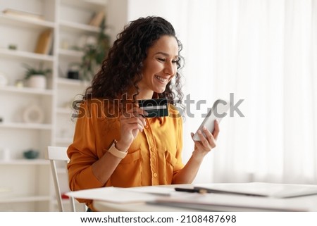Mobile Shopping. Cheerful Arabic Woman Using Smartphone Shopping Online Holding Credit Card Making Payment Sitting At Desk At Home. Internet Banking Application And E-Commerce. Side View Royalty-Free Stock Photo #2108487698