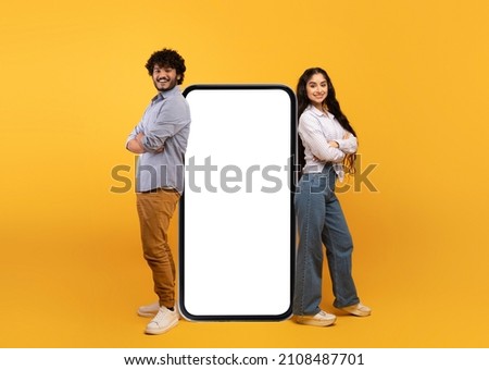 Millennial indian couple leaning on giant smartphone with mockup, promoting mobile application or website, advertising your service or product over yellow studio background Royalty-Free Stock Photo #2108487701