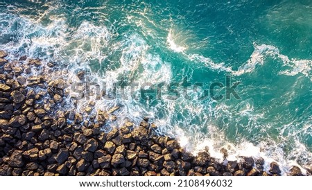 Beautiful drone shot of waves smoothly braking on shore with rocks at the sunset, with amazing blue and yellow colors. Top view.