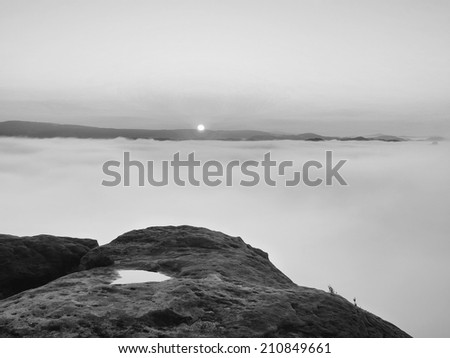 Dense fog in deep rocky valley full of heavy clouds. Sandstone peaks increased from foggy background, the Sun above. Black and white picture.