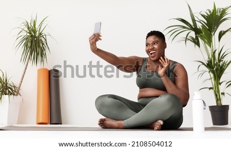 happy pretty chubby young black lady in sportswear sitting on fitness mat, holding smartphone and waving at gadget screen, taking selfie while doing workout at fitness studio, panorama, copy space