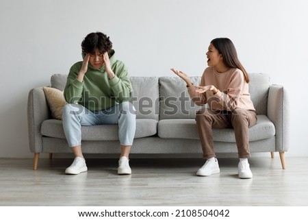 Angry millennial Asian wife yelling at her husband, man suffering from stress, sitting on sofa in living room, free space. Relationship problems, crisis, divorce and breakup concept Royalty-Free Stock Photo #2108504042