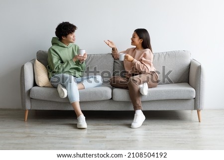 Young Asian boyfriend and girlfriend drinking coffee and chatting while sitting on sofa at home. Affectionate millennial couple enjoying hot drink, having conversation, spending time together Royalty-Free Stock Photo #2108504192