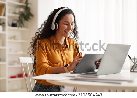 Cheerful Lady Wearing Headphones Using Digital Tablet And Laptop Computer Working Online Sitting At Desk In Modern Office. Female Entrepreneurship, Gadgets And Technology Concept. Side View Royalty-Free Stock Photo #2108504243