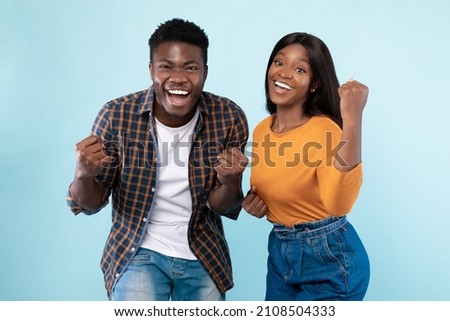 Wow, Yes. Portrait of overjoyed young black man and woman cheering and shaking clenched fists, looking at camera. Happy couple celebrating win posing standing isolated over blue studio background wall