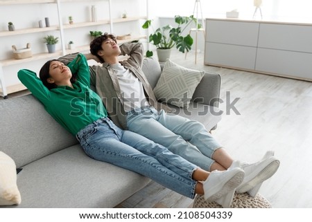 Calm young Asian couple relaxing on couch with their hands behind heads at home, blank space. Carefree girlfriend and boyfriend chilling on sofa, enjoying peaceful weekend morning Royalty-Free Stock Photo #2108504339