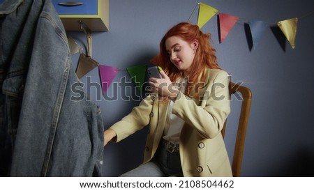 A beautiful red-haired girl paints with multicolored paints on clothes. The girl takes pictures of the finished work on her phone. Handmade work