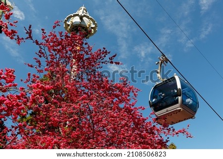 Tourists riding a gondola of the cable car over fiery cherry blossom trees (Sakura) and the Big Drop towering against blue sky, in Formosan Aboriginal Cultural Village, in Yuchi, Nantou County, Taiwan Royalty-Free Stock Photo #2108506823