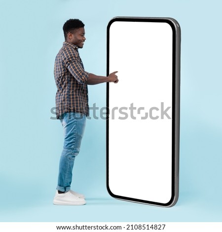 Side View Of Black Guy Using Big Cellphone With Blank White Screen Touching Huge Display Panel With Finger, Cheerful Man Standing On Blue Background, Ordering Food Delivery, Mock Up. Full Body Length Royalty-Free Stock Photo #2108514827