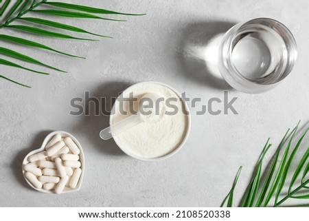 Collagen powder in a bowl, collagen capsules and a glass of water on a gray background with palm leaves. A natural supplement. Fish based or plant based. Top view, flat lay. Royalty-Free Stock Photo #2108520338