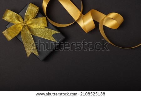 Black gift box with a golden ribbon and a large bow on a black  background. Place for the inscription. Boxing Day. Christmas holiday background.