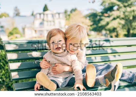 Outdoor portrait of two adorable siblings resting on the bench in sunny park, preschooler brother hugging sweet toddler sister Royalty-Free Stock Photo #2108563403
