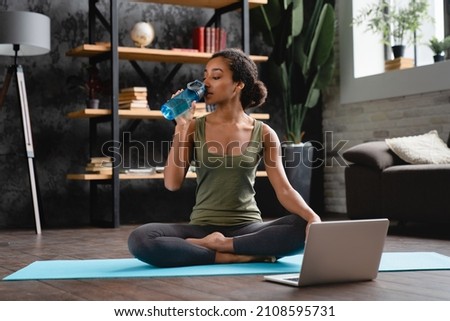 African fit young teenage girl woman in sporty clothes drinking water while sitting in yoga lotus position and watching online tutorial on laptop at home. Royalty-Free Stock Photo #2108595731