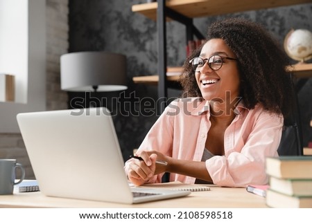 Cheerful laughing young african woman tutor freelancer student writing at copybook homework test, e-learning, working remotely, watching webinar on laptop