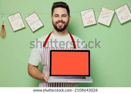 Young male chef confectioner baker man 20s in striped apron hold use work on laptop pc computer with blank screen workspace area isolated on plain pastel light green background Cooking food concept