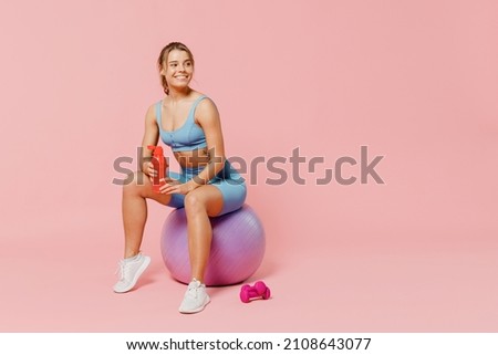 Full length young athletic fitness trainer instructor woman wear blue tracksuit spend time in home gym sit on fitball drink water isolated on pastel plain light pink background Workout sport concept