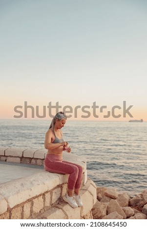 Full-length photo of asian lady in sportswear sitting on the seashore in the evening, opening a bottle of water